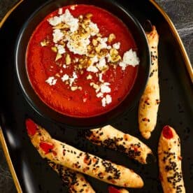 Halloween-themed Bloody Dip, a deep red roasted pepper and tomato spread topped with crumbled feta and chopped walnuts, served with witch fingers grissini