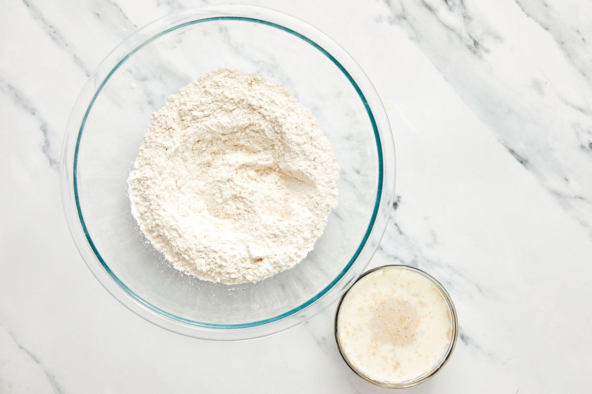 Dry ingredients for bao dough in a glass bowl with a small bowl of foamy yeast and warm milk on the side.