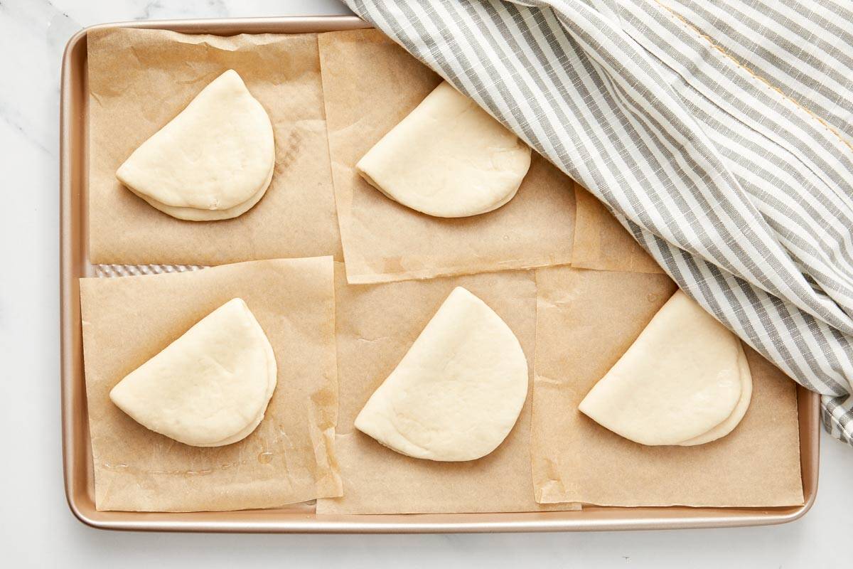 Folded bao buns proofing on parchment paper, covered lightly with a tea towel.