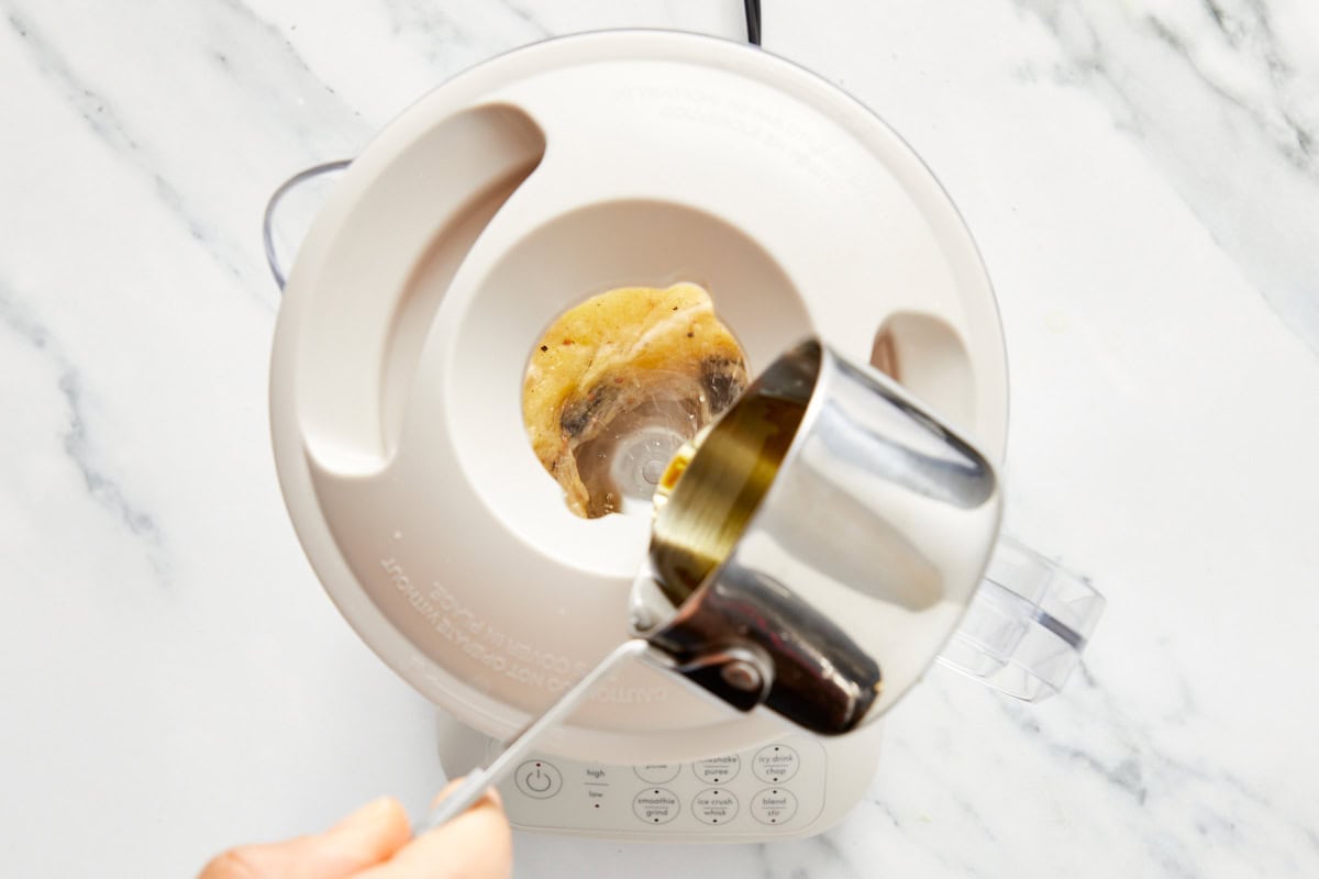 Olive oil being poured into a food processor with egg, black garlic, and mustard for making mayo.