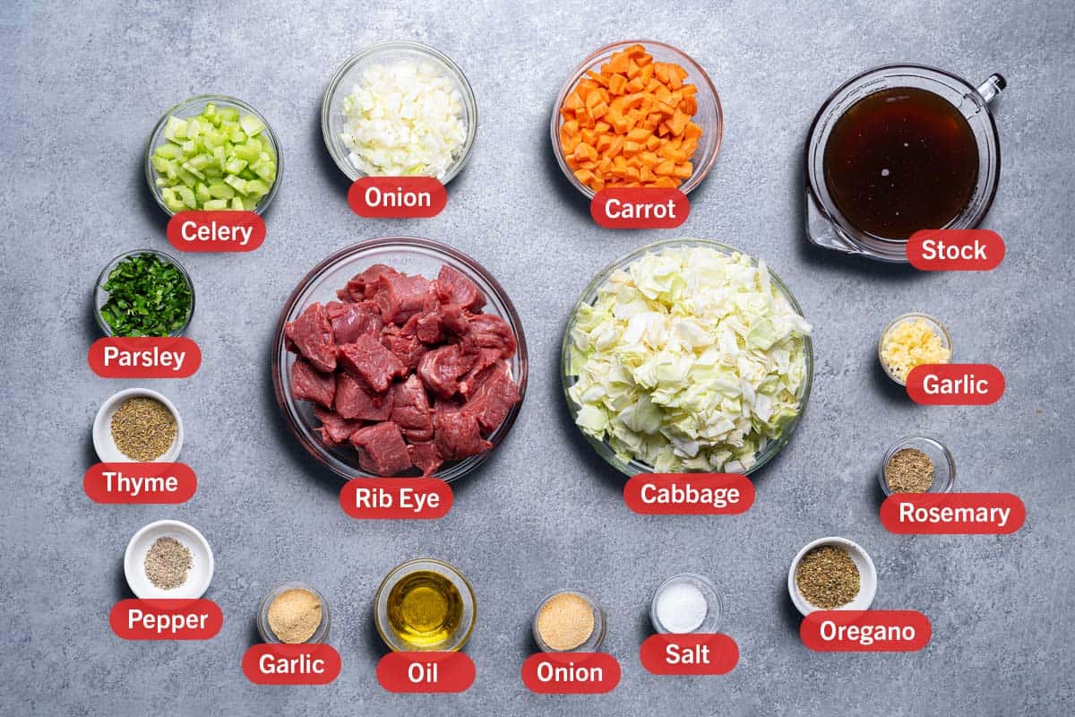 Grey board background with ingredients laid out, celery, onion, carrot. stock, parsley, rib eye, cabbage, garlic, thyme, pepper, garlic powder, oil, onion powder, salt, oregano, rosemary