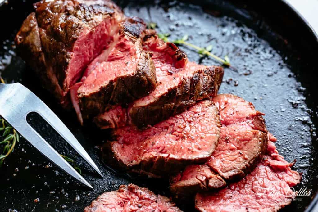 Roasted Beef in a black skillet cut into slices cooked to medium rare with sprigs of fresh rosemary and a crack of pepper over the meat.