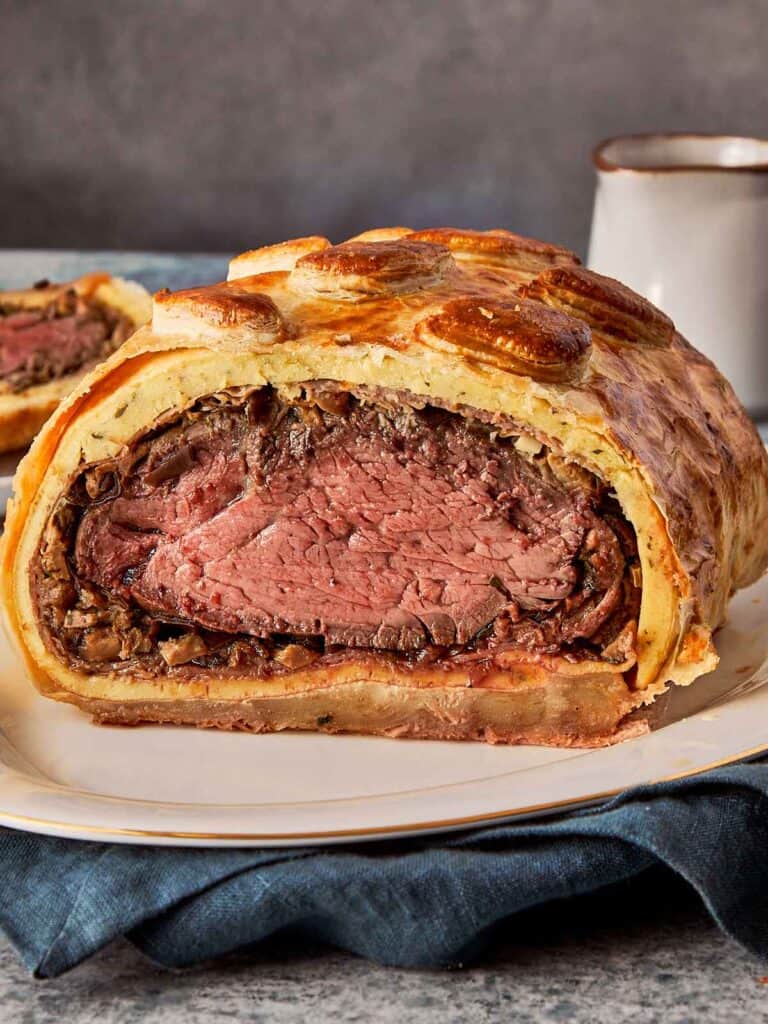 good beef wellington on a plate with cut in half exposing the pink meat inside covered with a good pastry