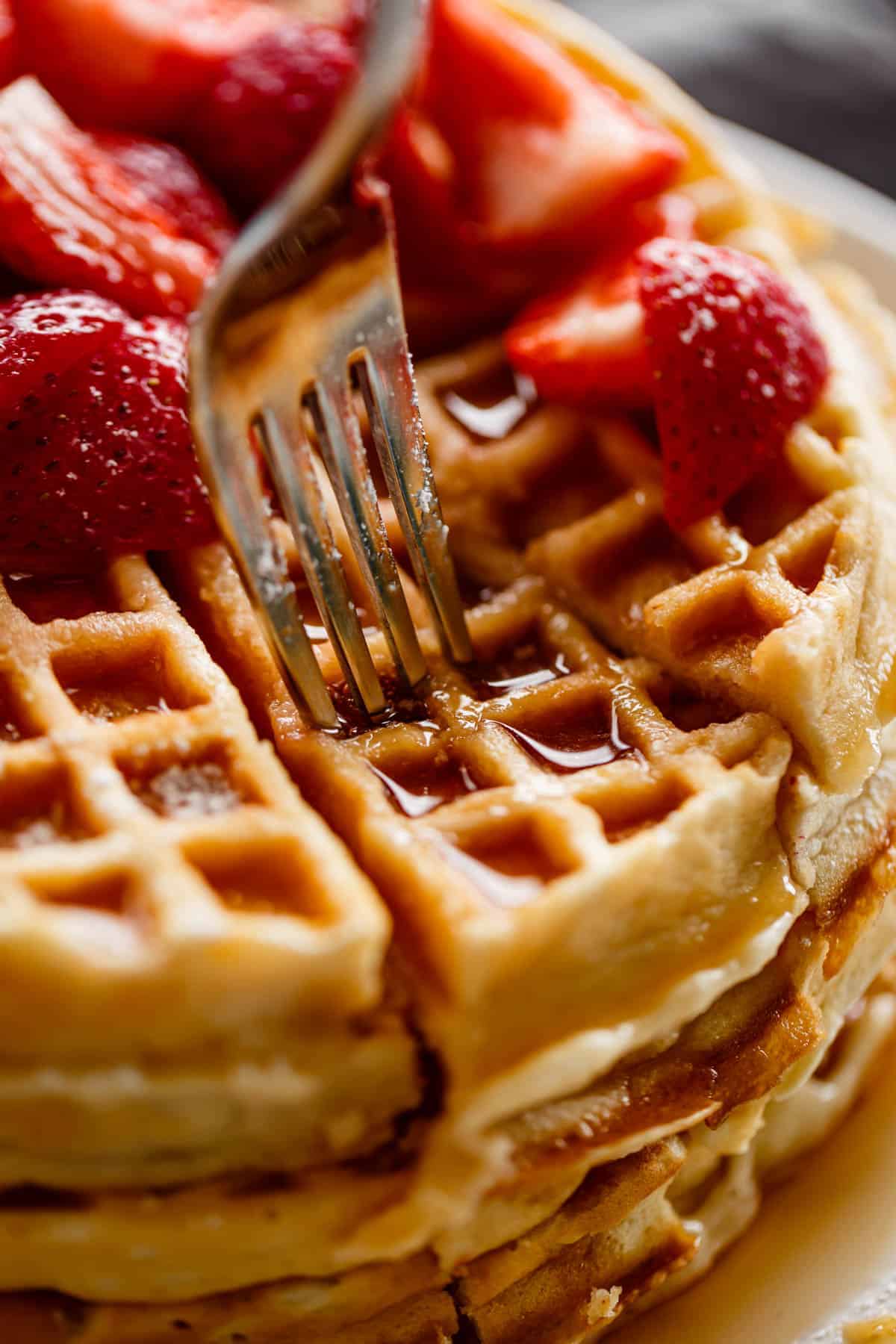 Close up image of fork piercing a stack of waffles, drenched in maple syrup and covered with chopped strawberries.