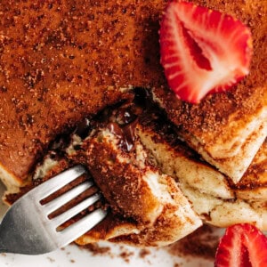 top view of a pancake stack, decorated in cinnamon sugar and fresh strawberries slice, fork opening a cut edge of stack to see the runny chocolate centre.