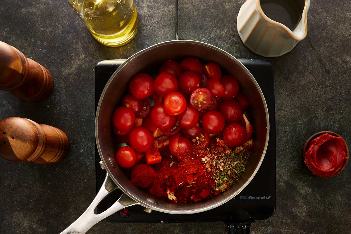 Overhead shot of cherry tomatoes, diced red capsicum, garlic, paprika, and herbs in a saucepan with olive oil ready to cook.