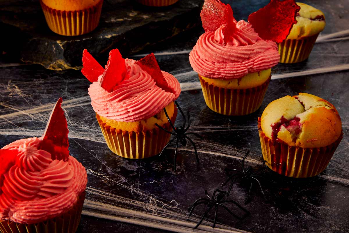 Group of raspberry muffins on a dark Halloween-themed surface, topped with pink frosting and candy glass shards.