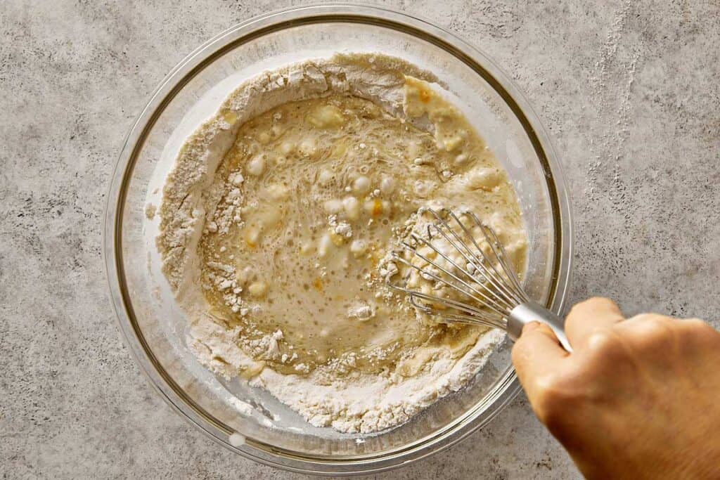Whisking the pancake batter as the wet and dry ingredients are gently combined in a glass bowl, leaving a few lumps for fluffy blueberry chocolate chip pancakes.
