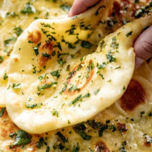 Holding a Garlic Naan Bread on a plate with a pile of naan. Garnished with fresh chopped cilantro.