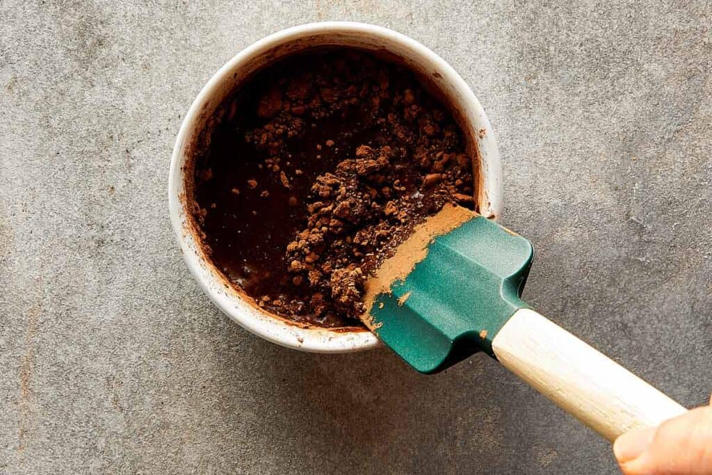 a small ceramic bowl sitting on a bench top with a mixing spatula with coffee and liquid inside.