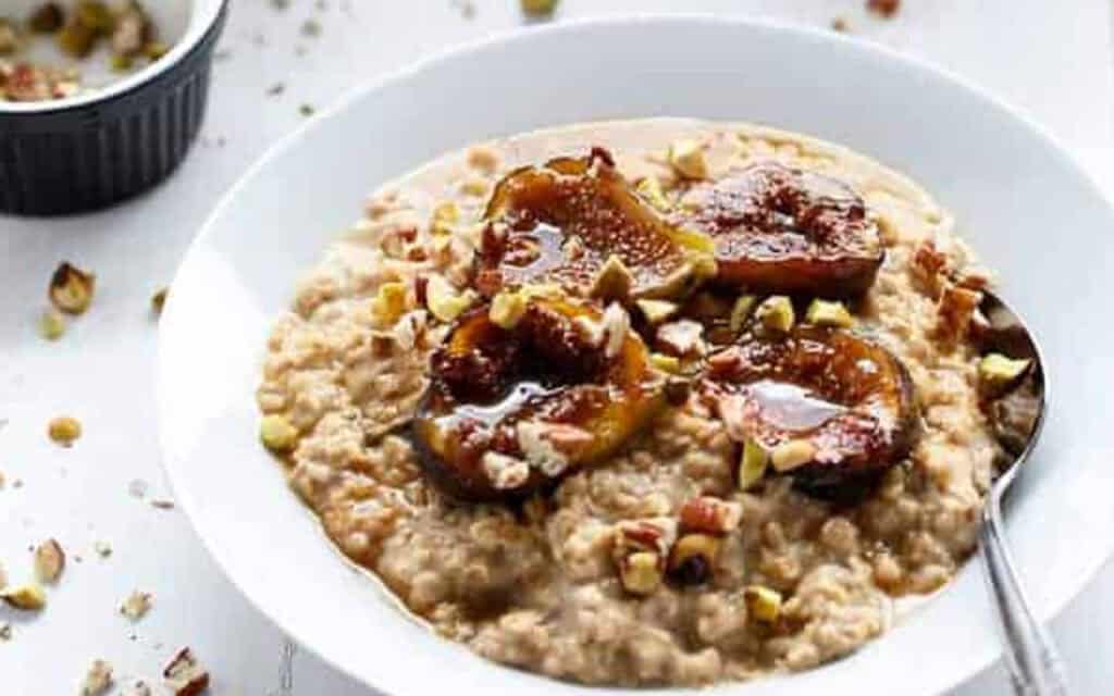 a good full bowl of porridge with caramelized figs on top and a spoon on the side, the table has a few scattered crushed pistachios.