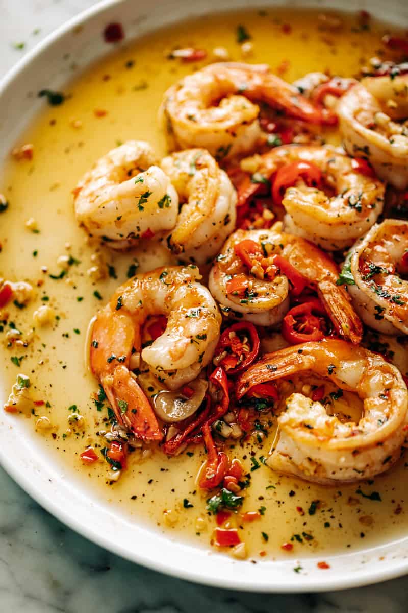 On a white plate, a serving of the chilli garlic butter shrimp in a bed of sauce with pieces of red chilli, green parsley.