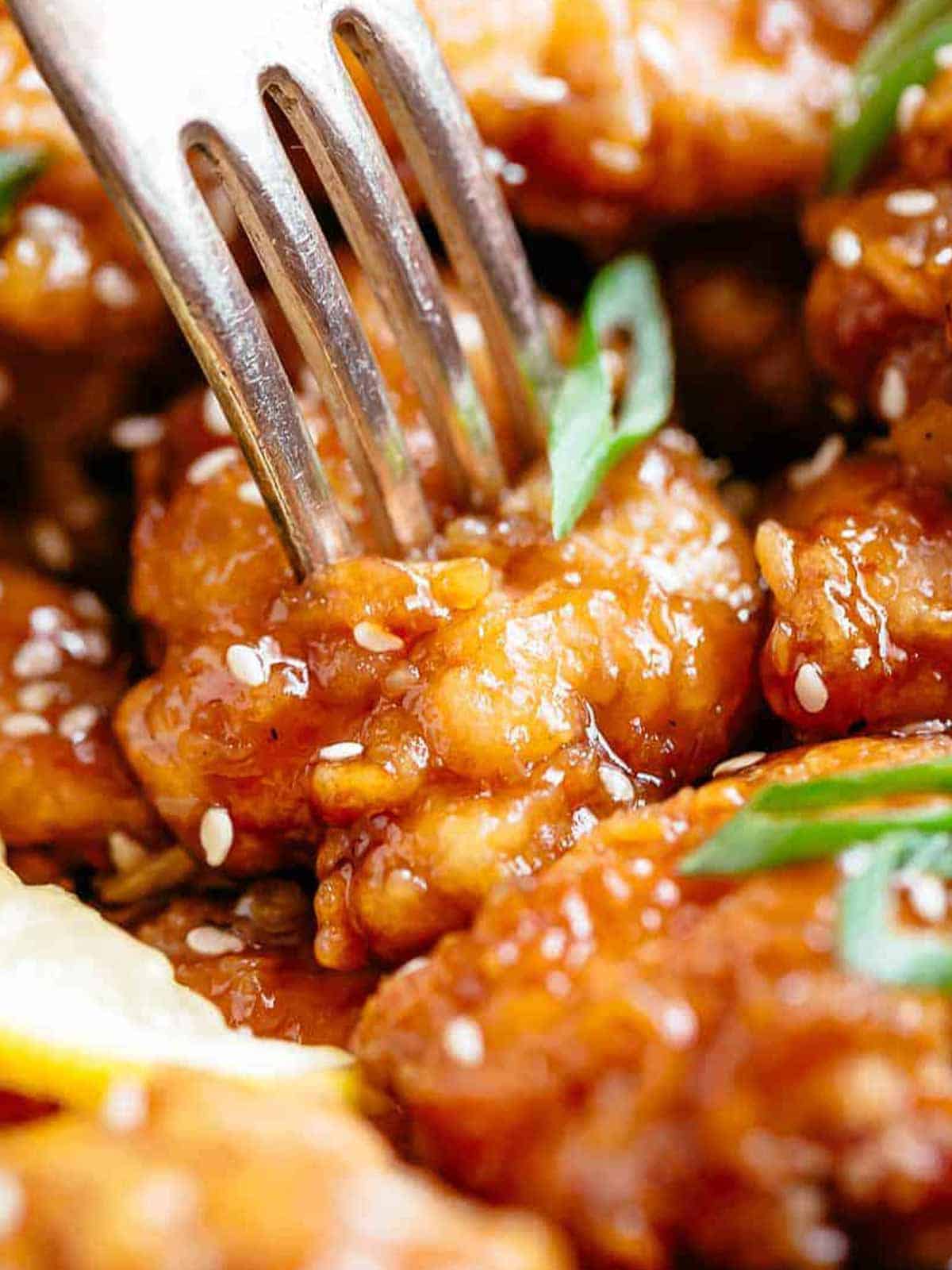 Close up of fork penetrating a crispy lemon chicken bite. The chicken is covered in sesame seeds and sliced spring onion. Looks good