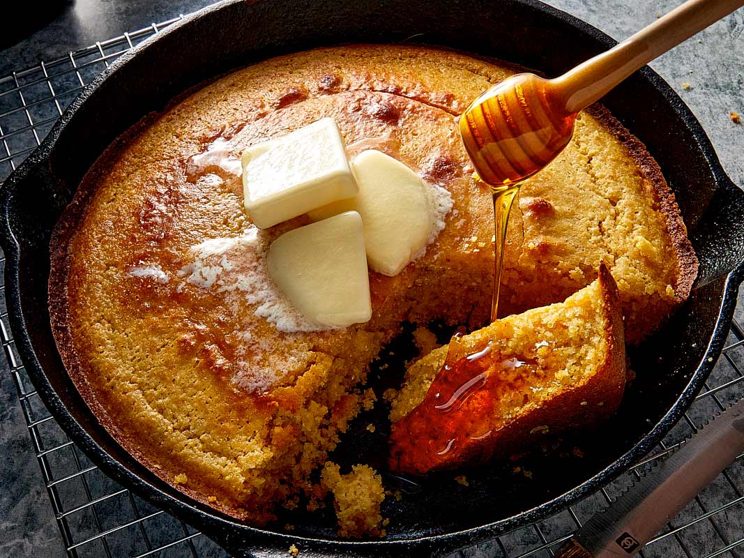 A freshly baked golden cornbread in a black cast iron skillet resting on a wire cooling rack. The cornbread has a slice cut and slightly pulled away, revealing a fluffy interior. Three pats of butter are melting on top of the warm cornbread, and honey is being drizzled onto the cut slice from a wooden honey dipper.