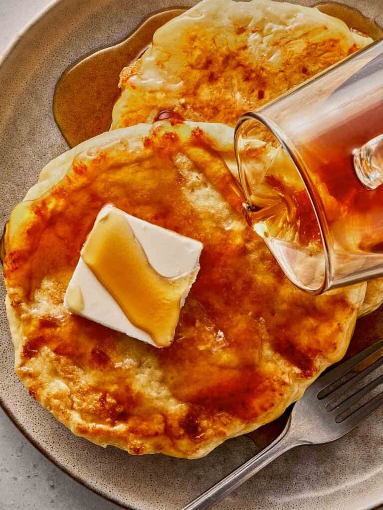 Cottage cheese pancakes on a plate with butter melting on top, drizzled with maple syrup as it's being poured, showing a soft texture and golden surface.