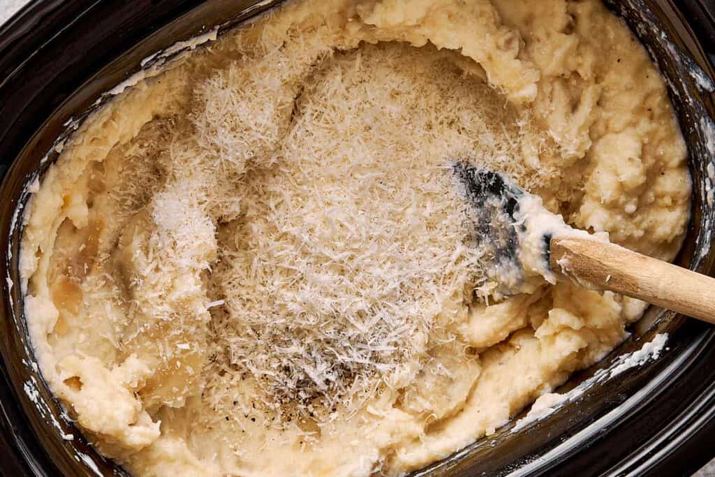 a top down view of mashed potatoes creamy stirred by a wooden spoon in a slow cooker with parmesan cheese on top