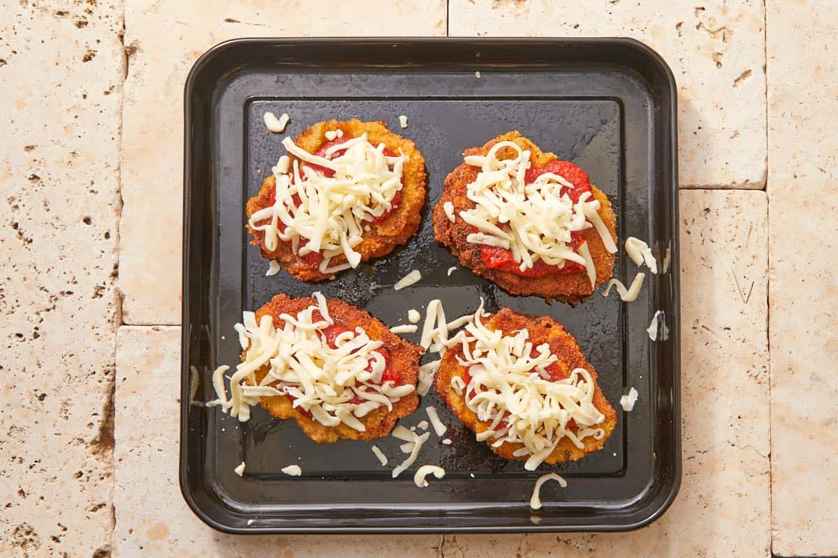 Beef schnitzels topped with sauce and shredded mozzarella, neatly placed on a black baking tray.