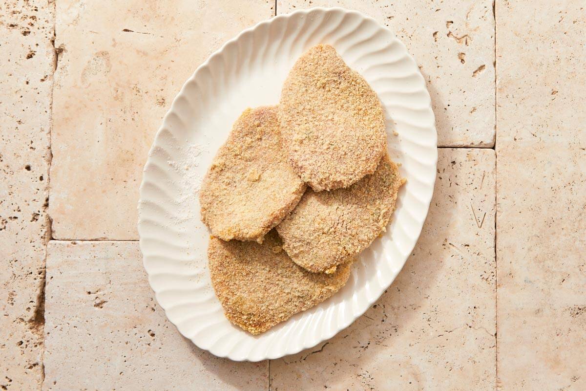 Four breaded schnitzels stacked on a white plate after being coated.