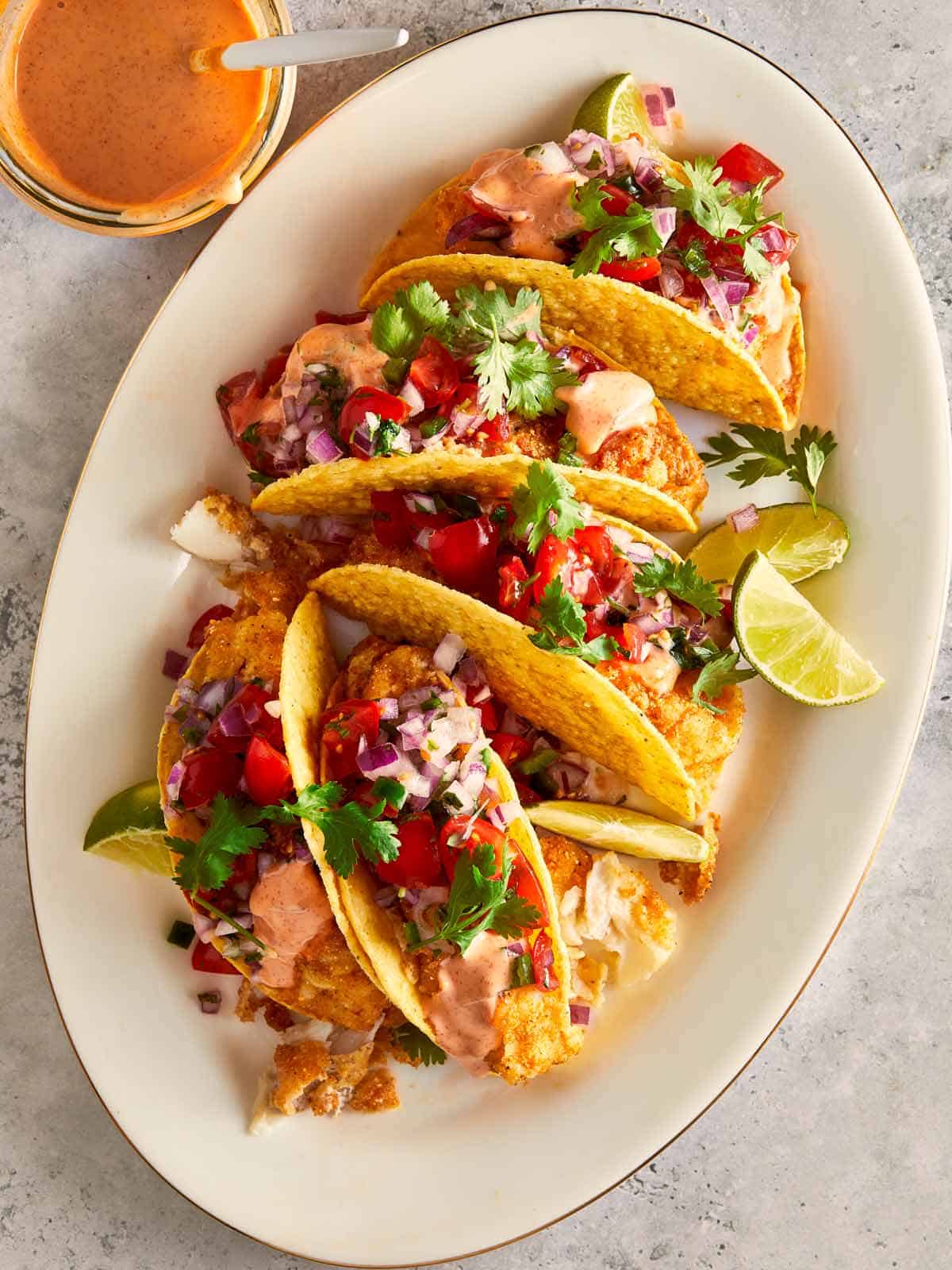 Plate of crispy fish tacos with pico de gallo, spicy mayo, cilantro, and lime wedges