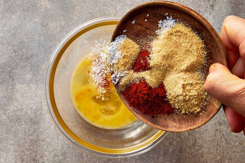 a top down view of a bowl filled with spices being poured into a glass mixing bowl that is filled with mixed egg wash