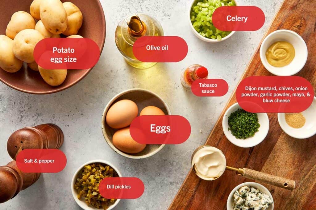 Ingredients for deviled potatoes including baby potatoes, eggs, mayo, Dijon mustard, dill pickles, celery, chives, blue cheese, olive oil, and seasonings arranged on a kitchen surface