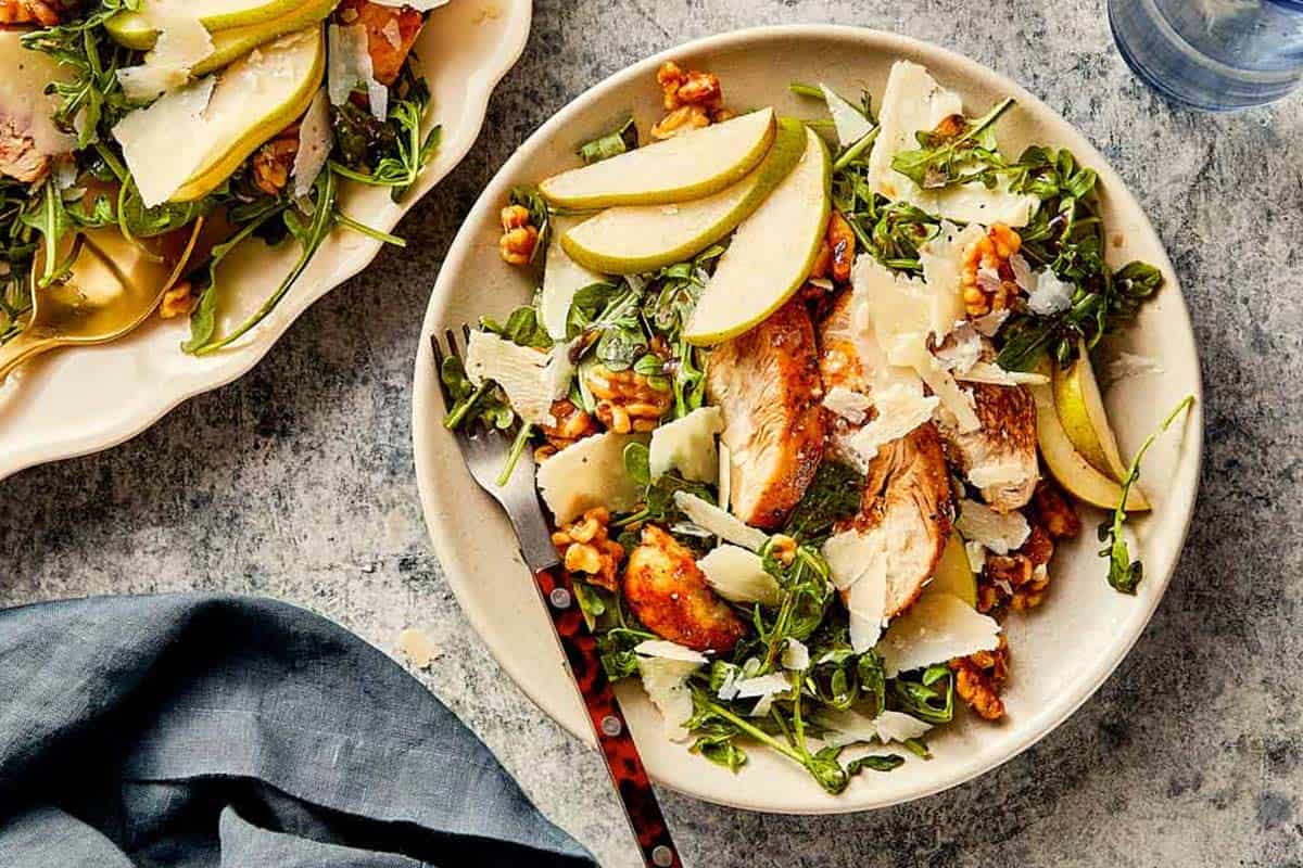 Serving of arugula salad with grilled chicken, pears, walnuts, and parmesan on a ceramic plate.