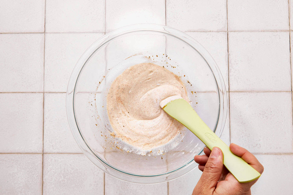 Hand mixing a creamy creek yogurt with spices in a glass bowl using a green spatula.