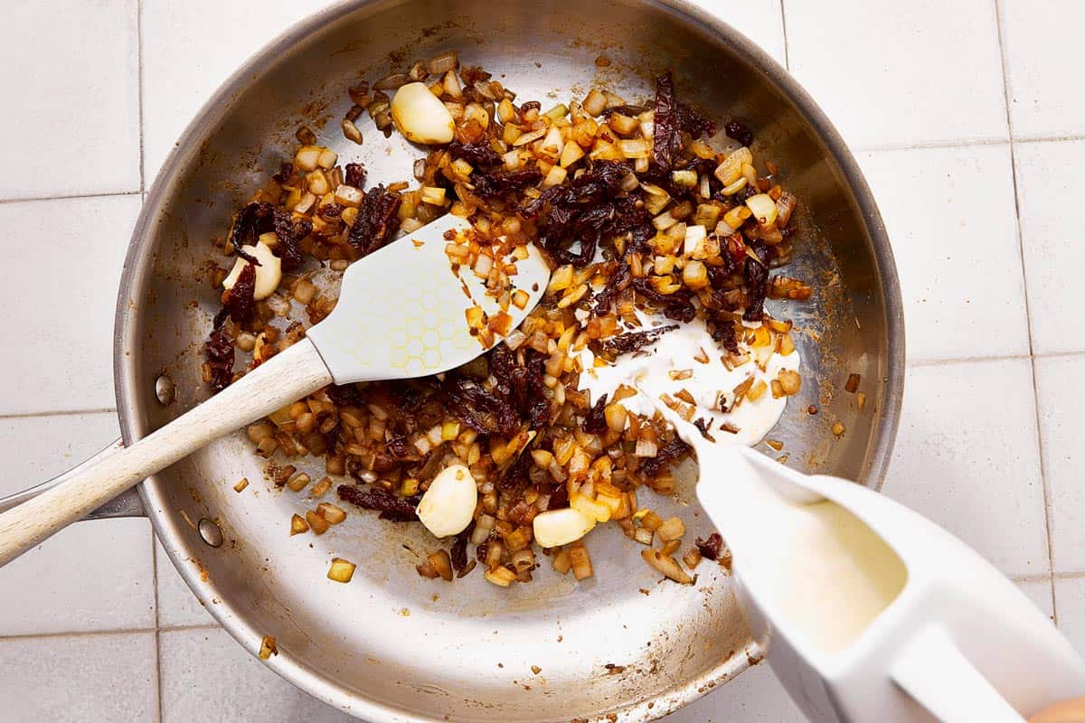 Pouring cream into a skillet with sautéed onions, garlic, and sun-dried tomatoes, while stirring with a spatula.