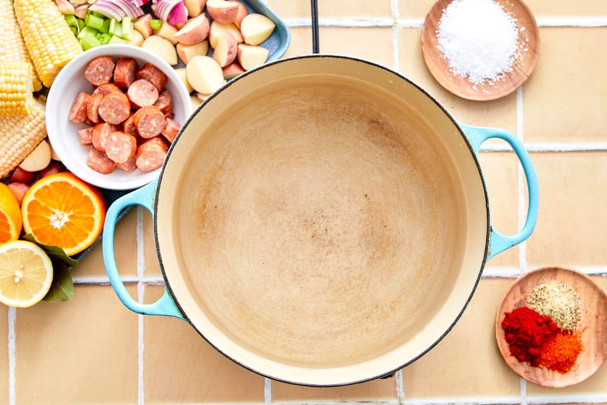 Empty pot surrounded by seafood boil ingredients including spices, garlic, onions, and citrus.