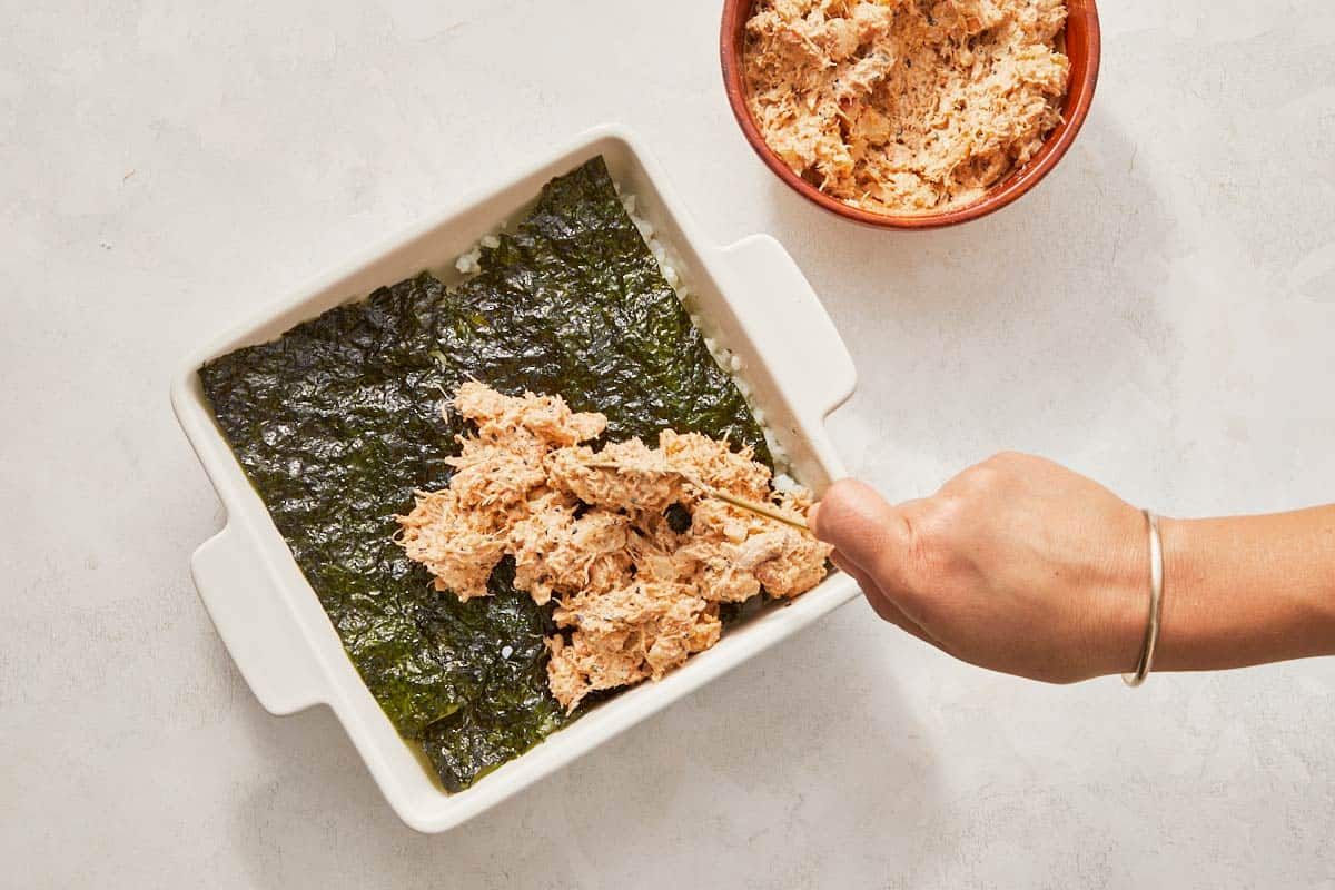 Spoon over a dish of food, preparing to add nori and a creamy seafood topping to the rice.