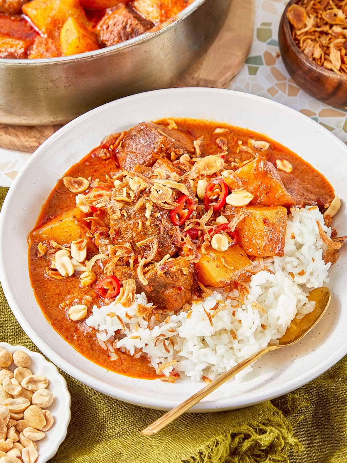 Plate of Massaman Beef Curry with potatoes and beef served over jasmine rice, topped with peanuts, red chili, and crispy shallots