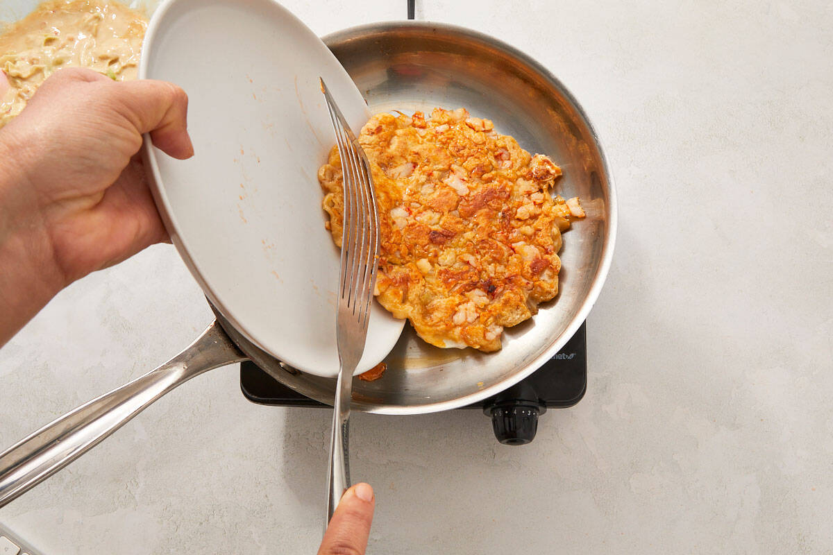 A spatula lifting the okonomiyaki as it's being flipped in the pan with a lid held to the side.