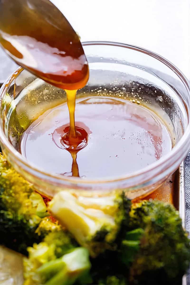 A metal spoon drizzling thick, glossy honey garlic sauce into a clear glass bowl, with fresh green broccoli florets visible on a baking sheet in the foreground.