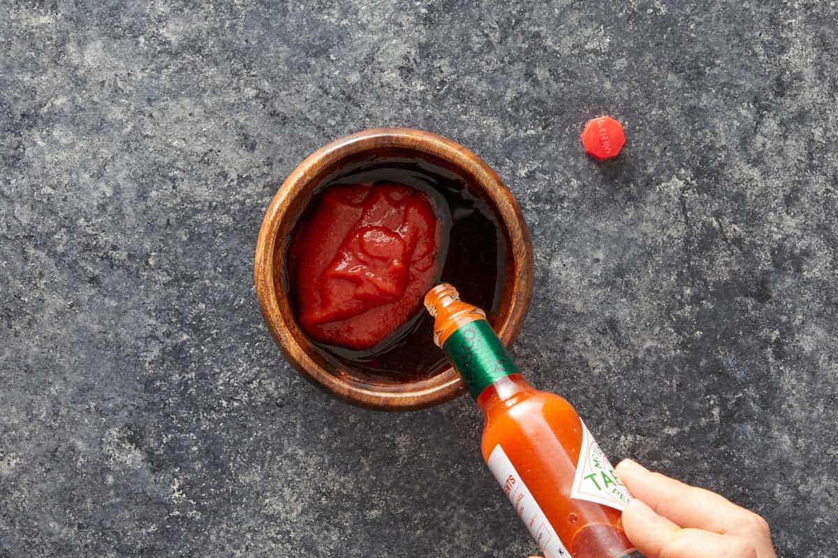 Hand pouring Tabasco sauce into a bowl of tomato sauce.