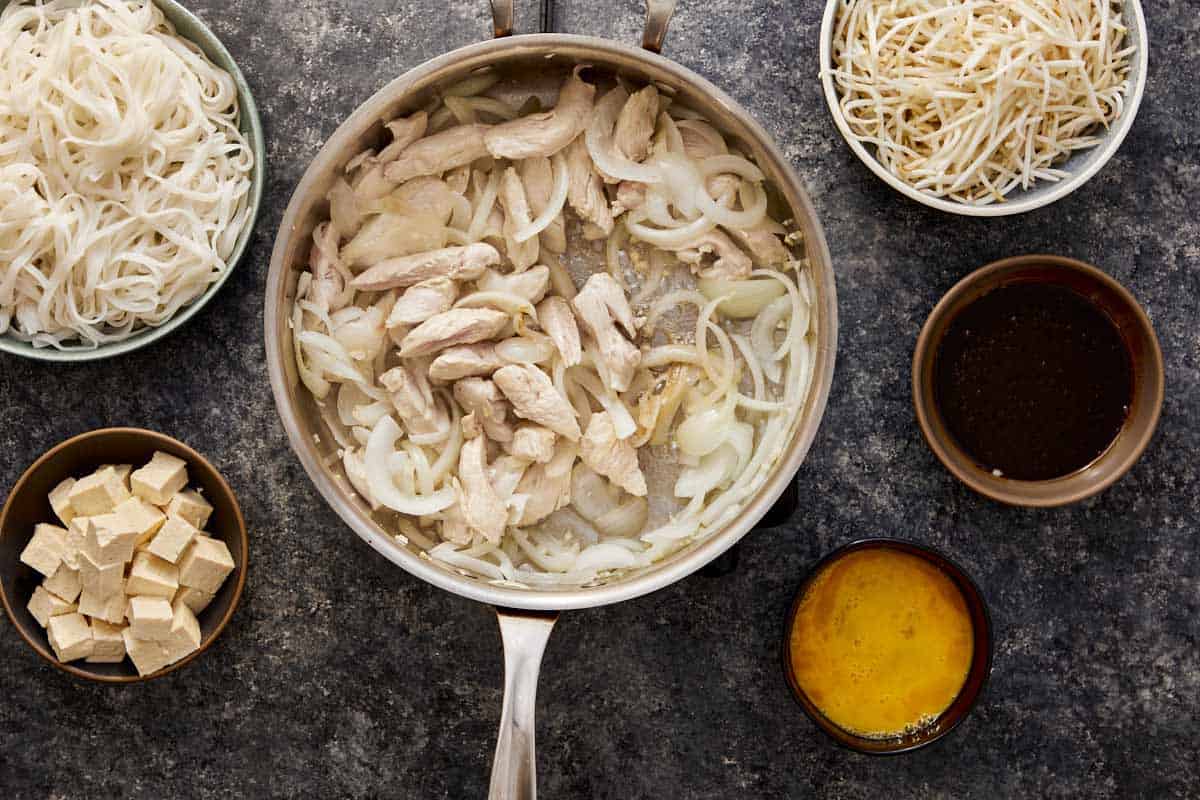Chicken strips cooking with onion and garlic in a pan, surrounded by Pad Thai ingredients.
