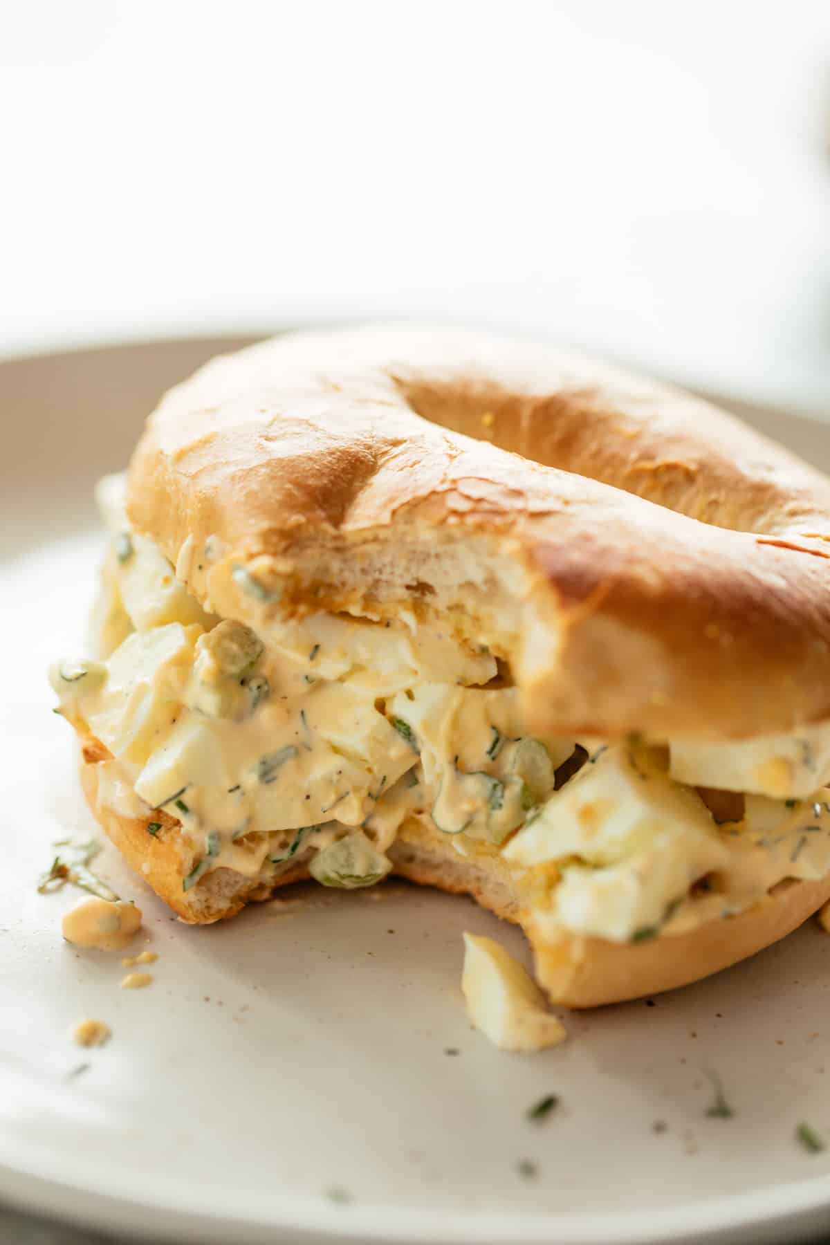good Egg Salad sandwiched in a bagel on a white plate sprinkled with fresh dill, chives and parsley.