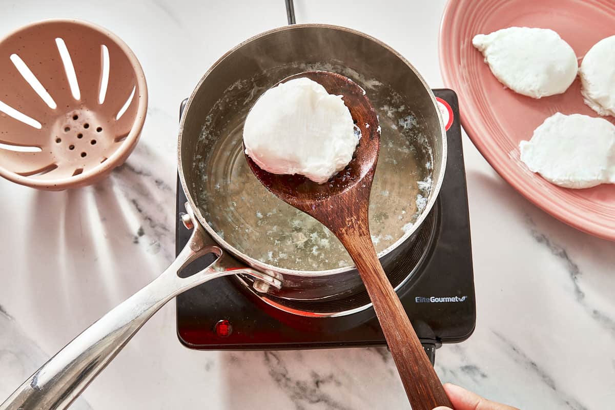 A poached egg being lifted from hot water with a slotted spoon, surrounded by perfectly cooked eggs on a plate.