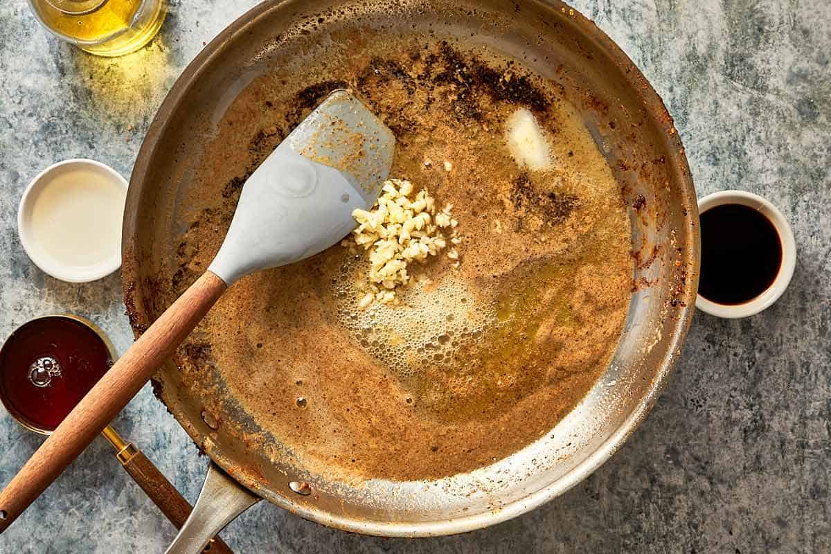a top down shot of a frying pan with butter melting inside and a spatula mixing spices the butter has melted and the garlic is frying in the centre of the pan