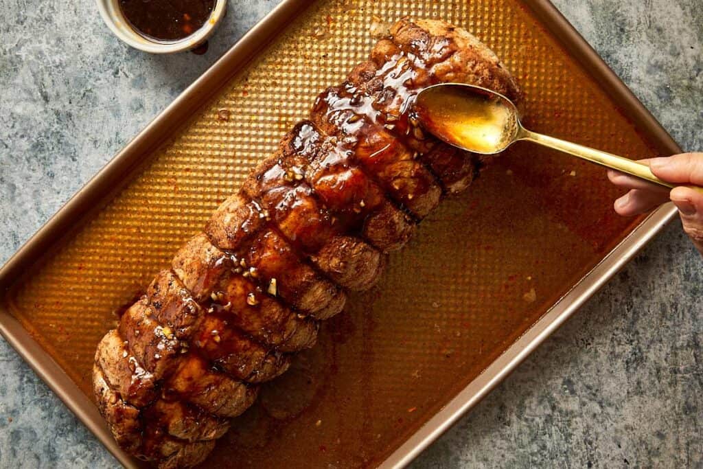 a long pork loin resting on a perforated oven rack ready for baking with a spoon of glaze covering the roast