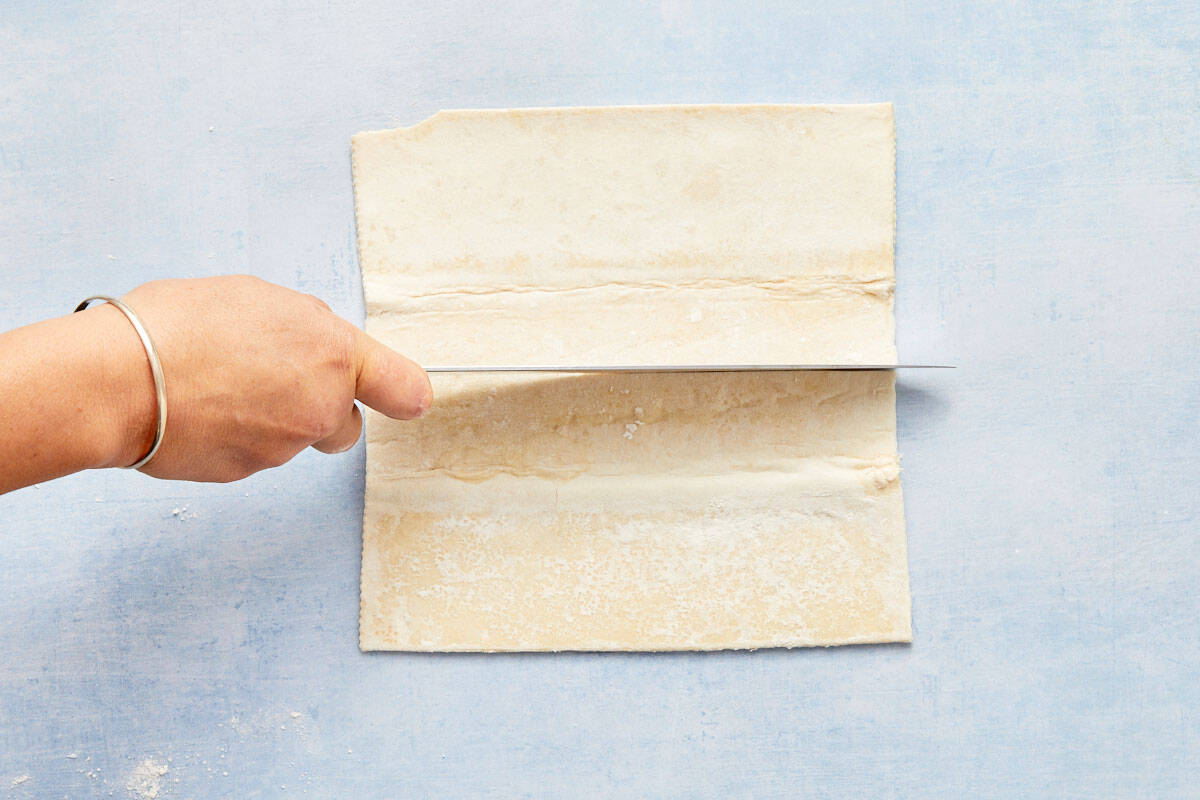 Puff pastry sheets being cut into rectangles on a light blue background.