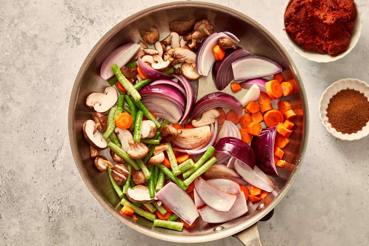 Sliced red onion, carrots, long beans, and shiitake mushrooms sizzling in a pan, the first step to build color and texture for the curry.