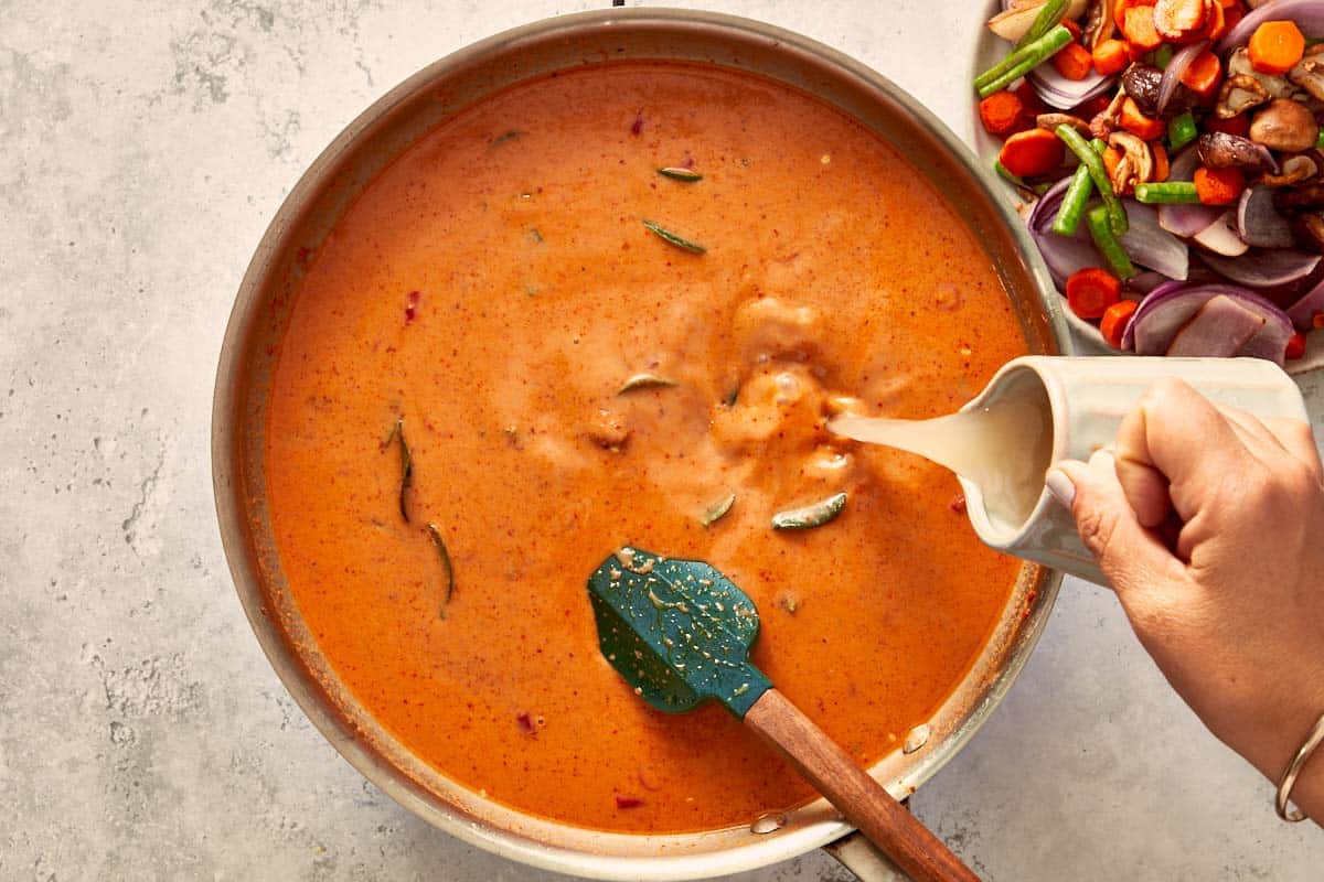 Coconut milk and chicken stock being poured into the curry base, creating a creamy orange sauce with floating lime leaves.