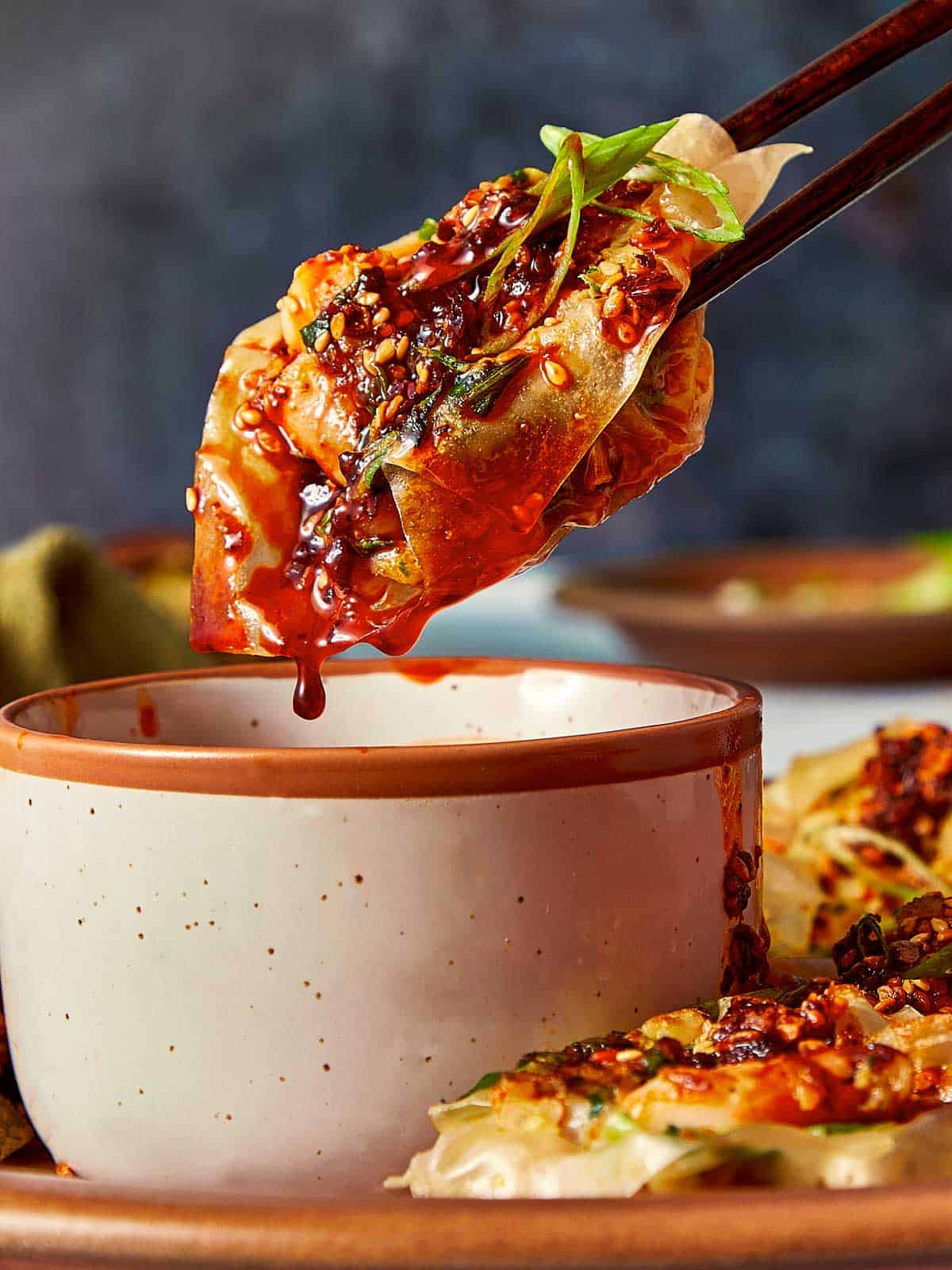 Close-up of a cooked shrimp dumpling held by chopsticks, dripping dark chili sauce into a small bowl, garnished with sesame seeds and sliced green onions.