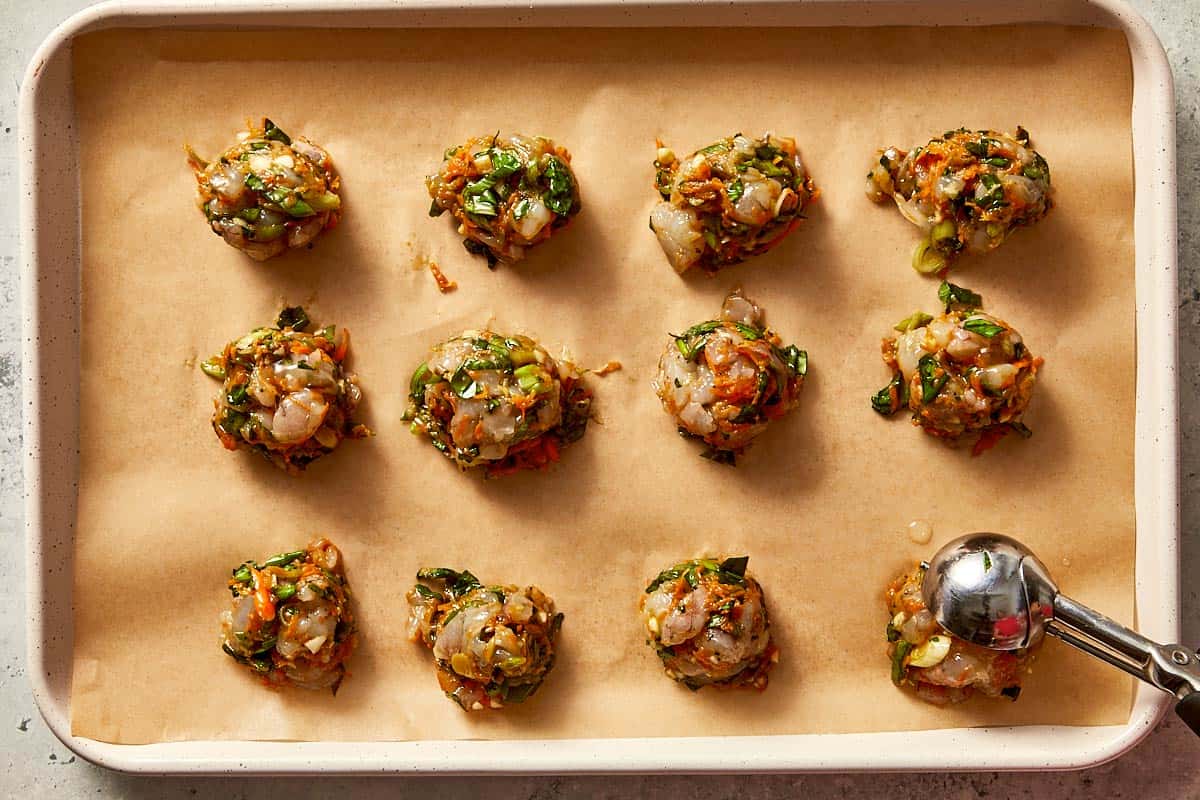 Raw shrimp dumpling filling scooped into small balls on a parchment-lined baking sheet, with a metal cookie scoop used for portioning.