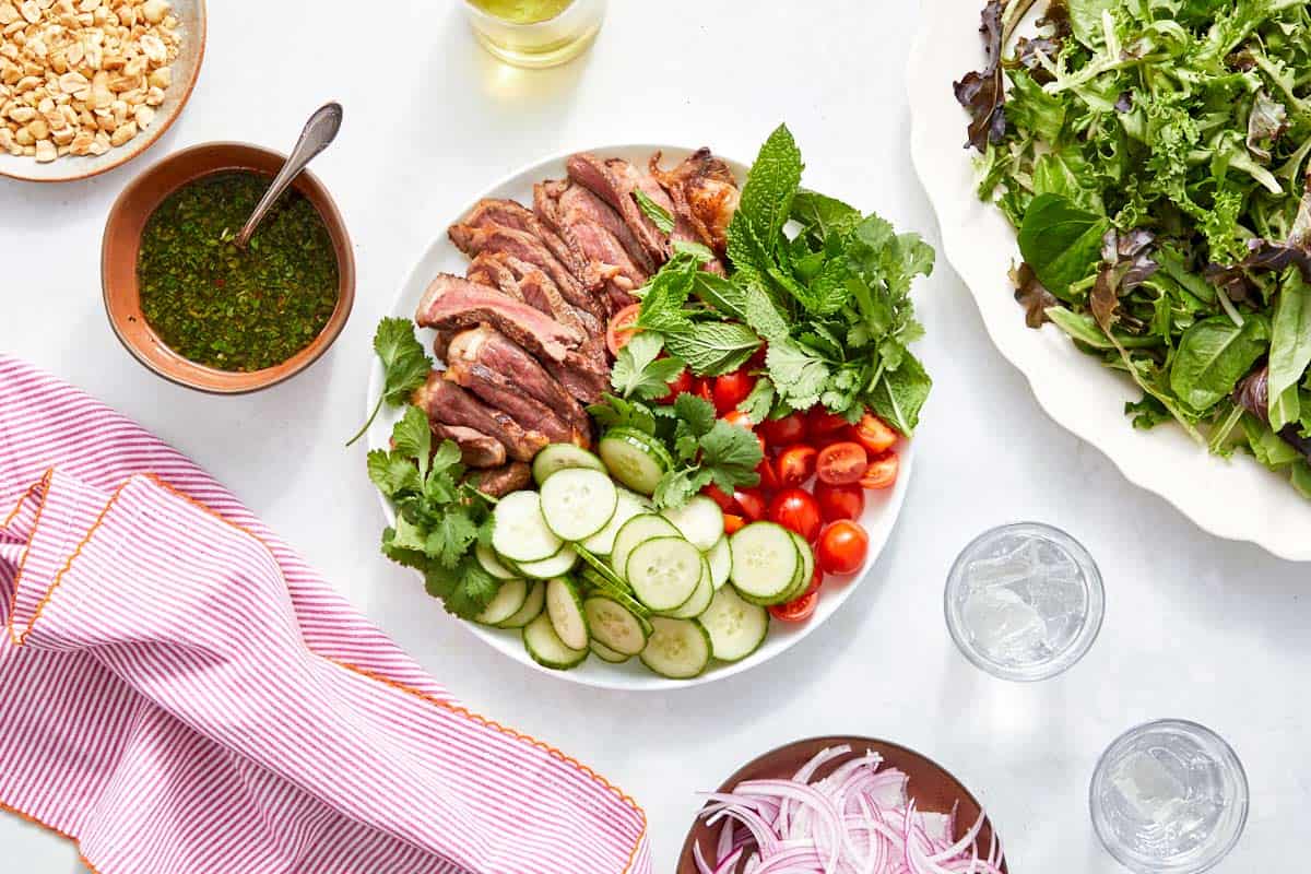 Salad components arranged, steak, cucumber, tomatoes, herbs, dressing.
