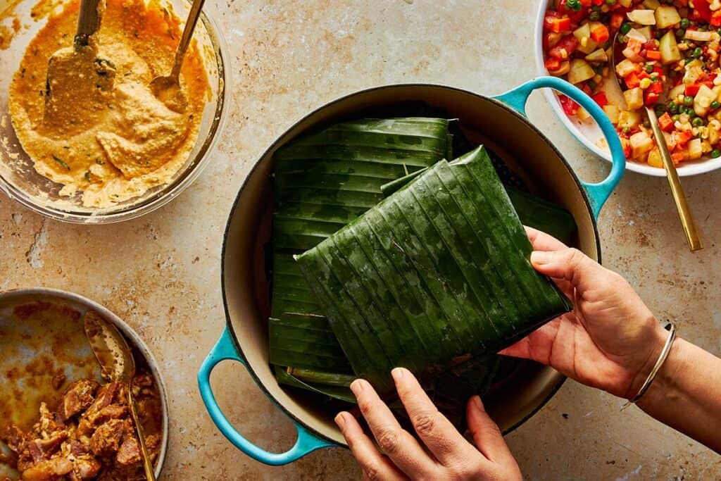 Overhead shot of hands placing a tamale wrapped in fresh green banana leaf into a teal Dutch oven for steaming