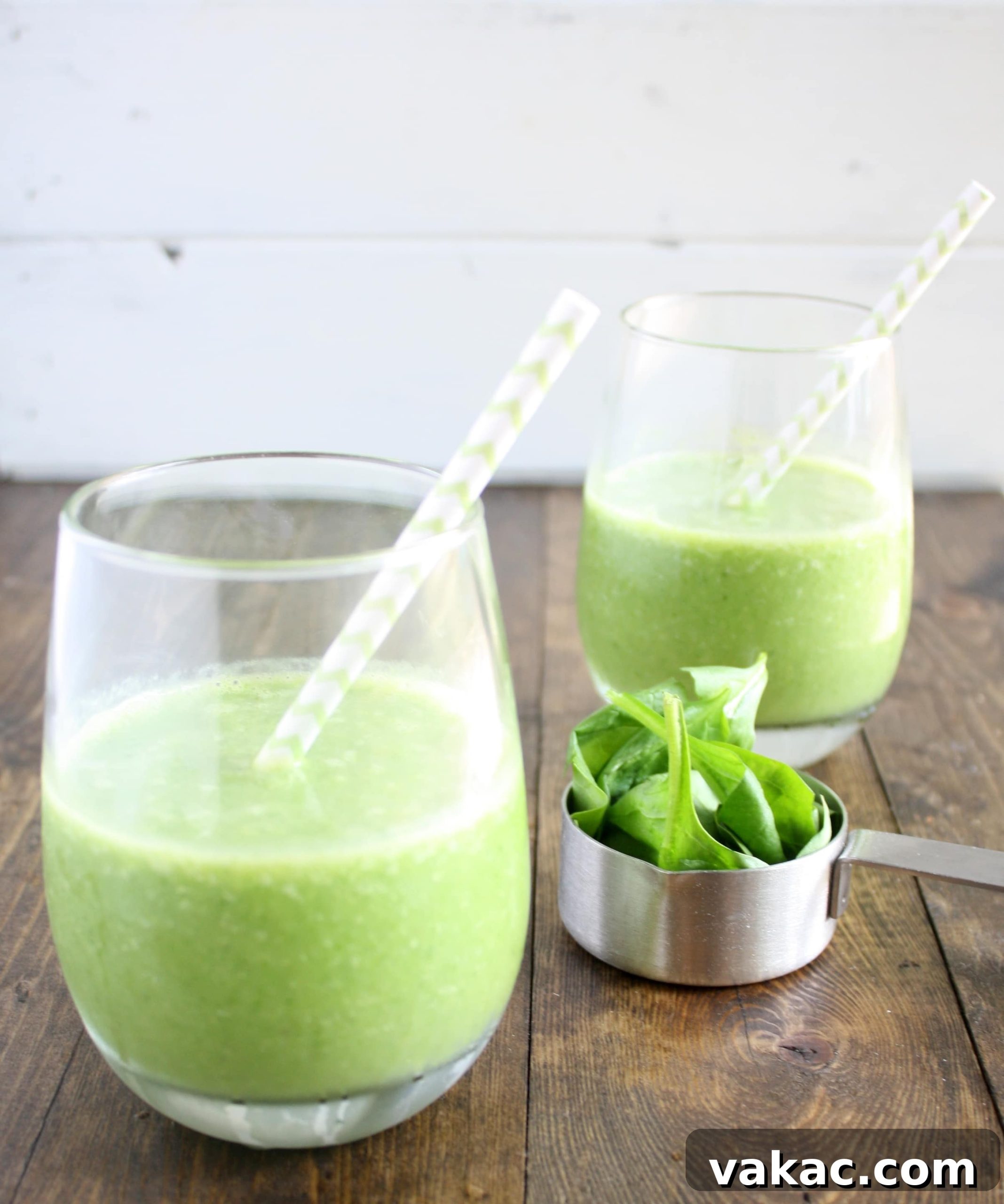 Rejuvenating Green Cleanse 4 Side view of two spinach banana pineapple fruit smoothies with straws. A measuring cup of fresh spinach rests between the glasses, highlighting the key ingredient.