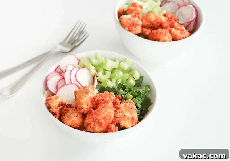 Two white bowls on a white surface with baked buffalo cauliflower, chives, celery, rice, and watermelon radish.