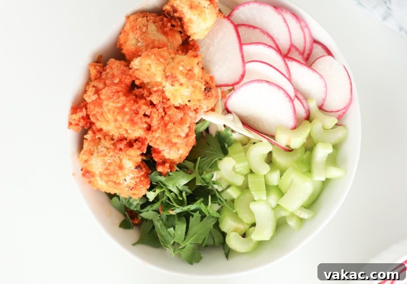 A white bowl on a white surface with baked buffalo cauliflower, greens, celery, rice, and watermelon radish.