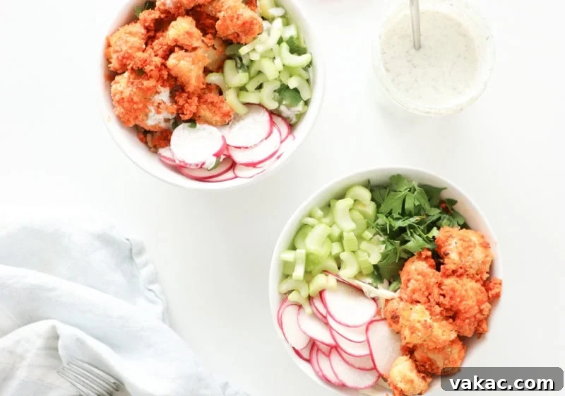 Two white bowls on a white surface with baked buffalo cauliflower, greens, and watermelon radish with a light blue kitchen towel and jar of homemade ranch dressing.